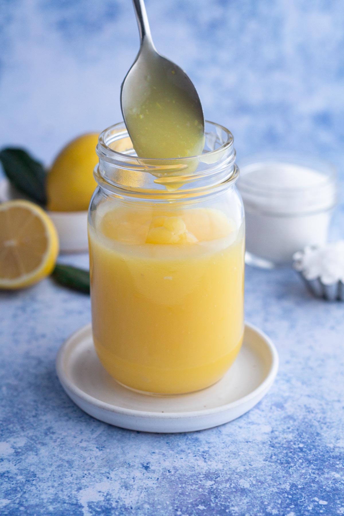 Spoon scooping lemon curd from a glass jar on a blue surface.