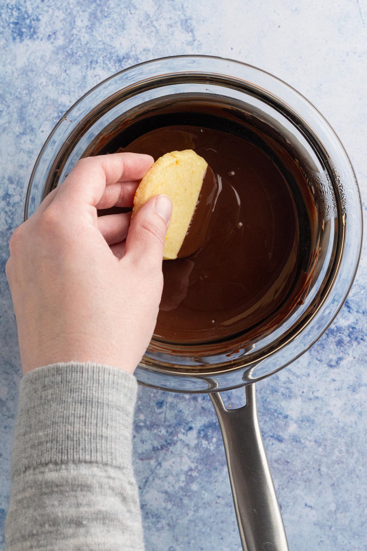 Hand dipping a shortbread cookie into melted chocolate in a double boiler.