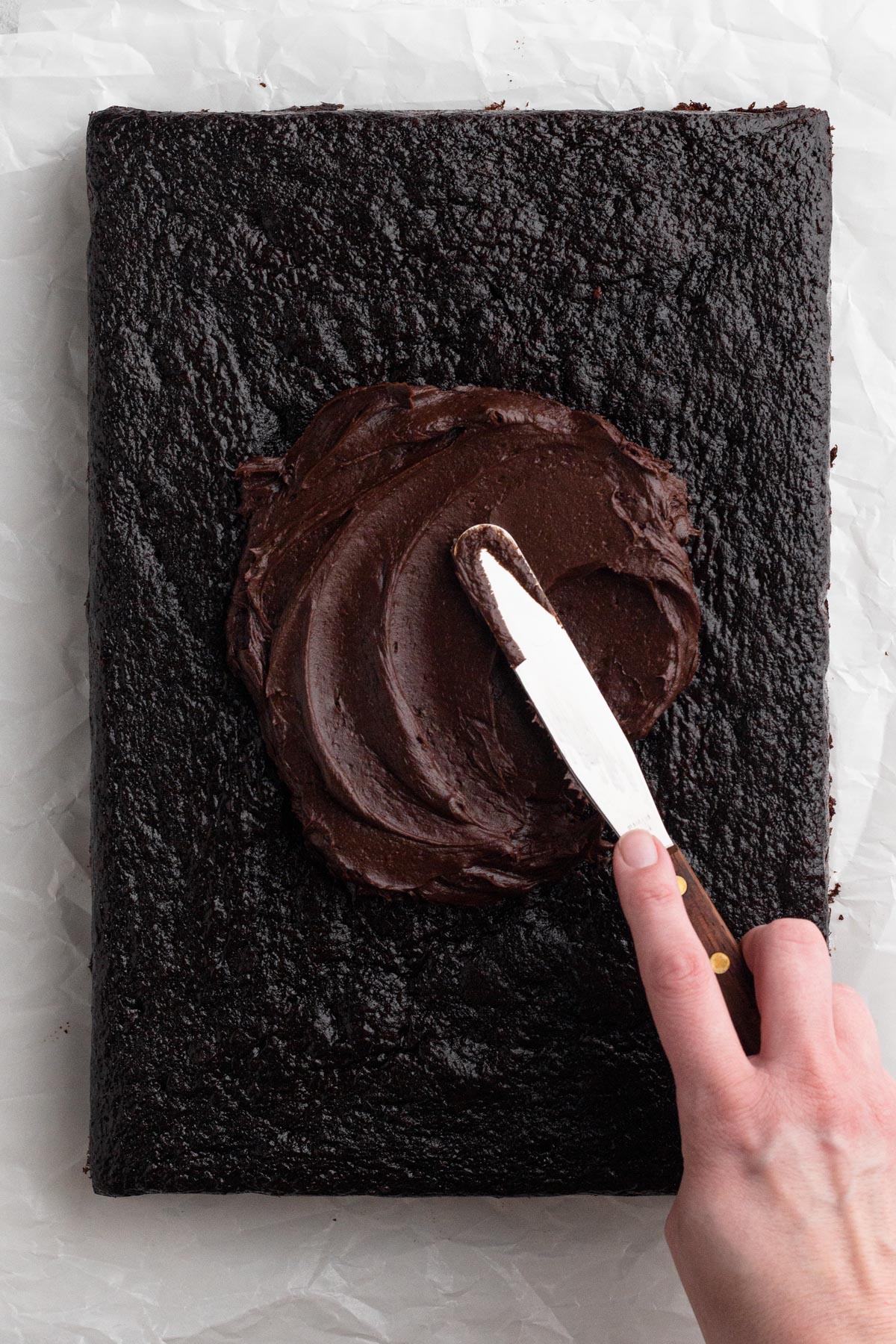 Overhead view of a hand using a spatula to spread chocolate frosting onto a chocolate sheet cake.