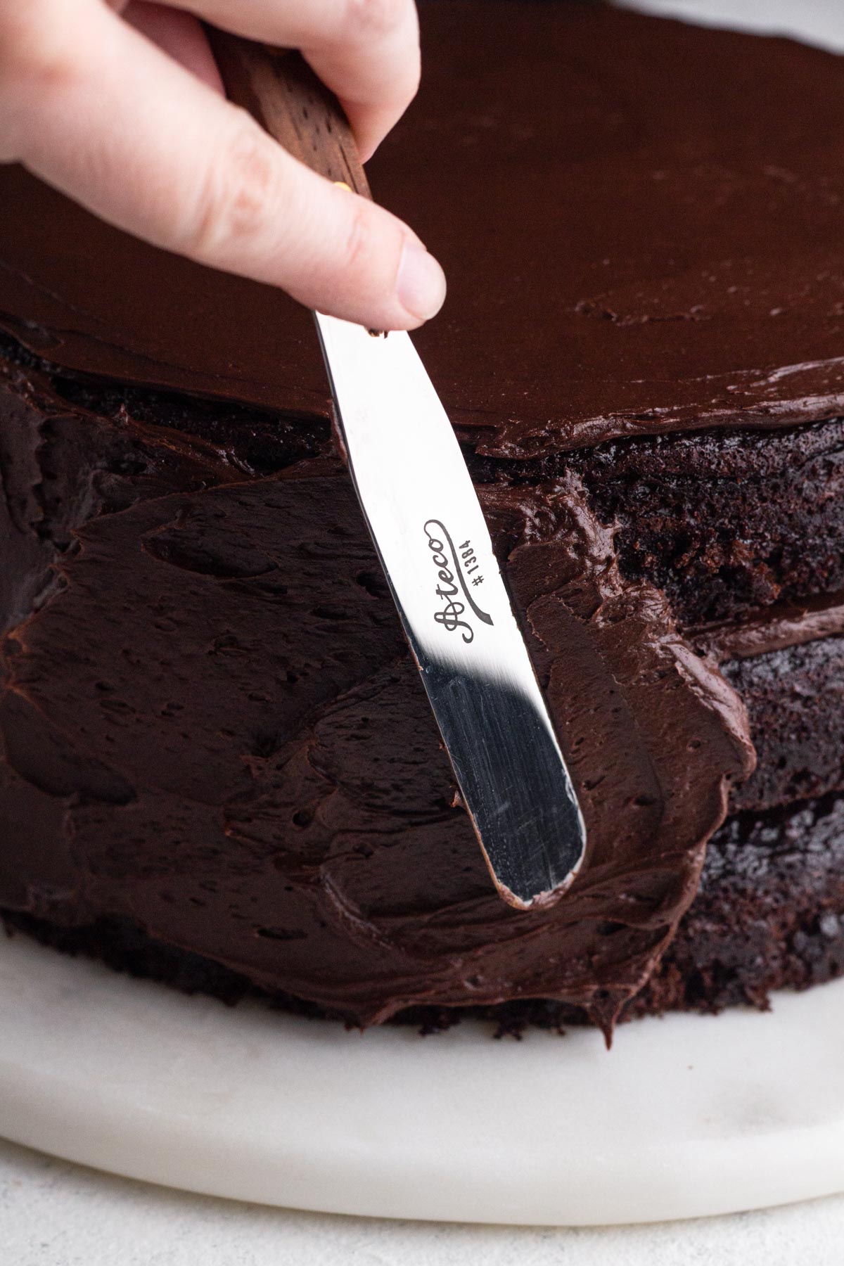 Side view of hand using a spatula to spread dark chocolate frosting along the side of a chocolate layer cake.