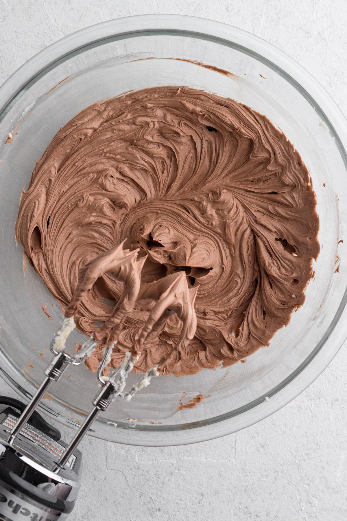 Beaten frosting in a glass mixing bowl with an electric hand mixer.
