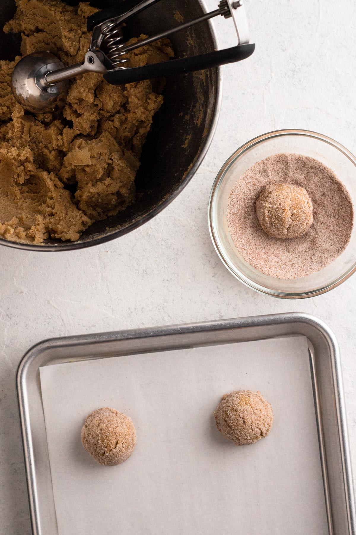 Bowl of cookie dough with cookie scoop and cookie dough balls rolled in sugar on a baking sheet lined with parchment paper.