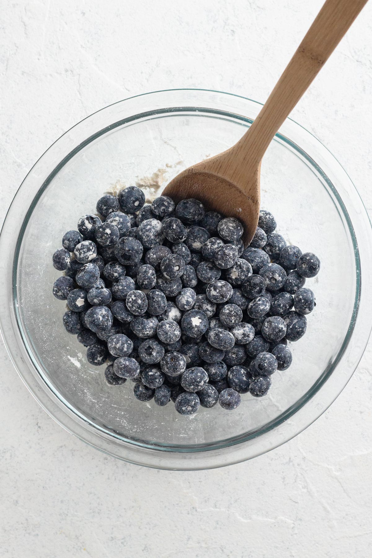 Fresh blueberries tossed in flour in a glass mixing bowl with a wooden spoon.