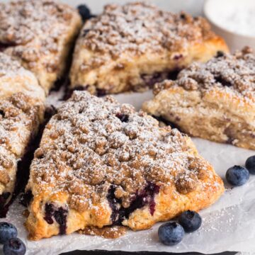 Blueberry scones surrounded by fresh blueberries and topped with streusel and powdered sugar.