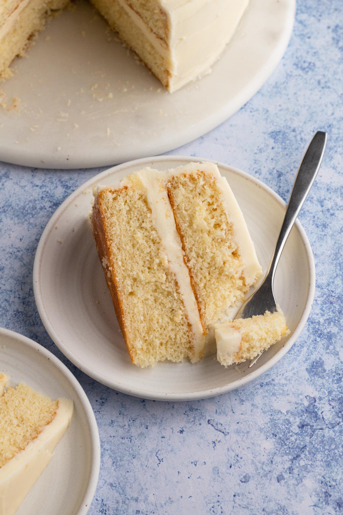 A slice of two-layer vanilla cake with vanilla frosting on a white plate next to the cake on a marble board.