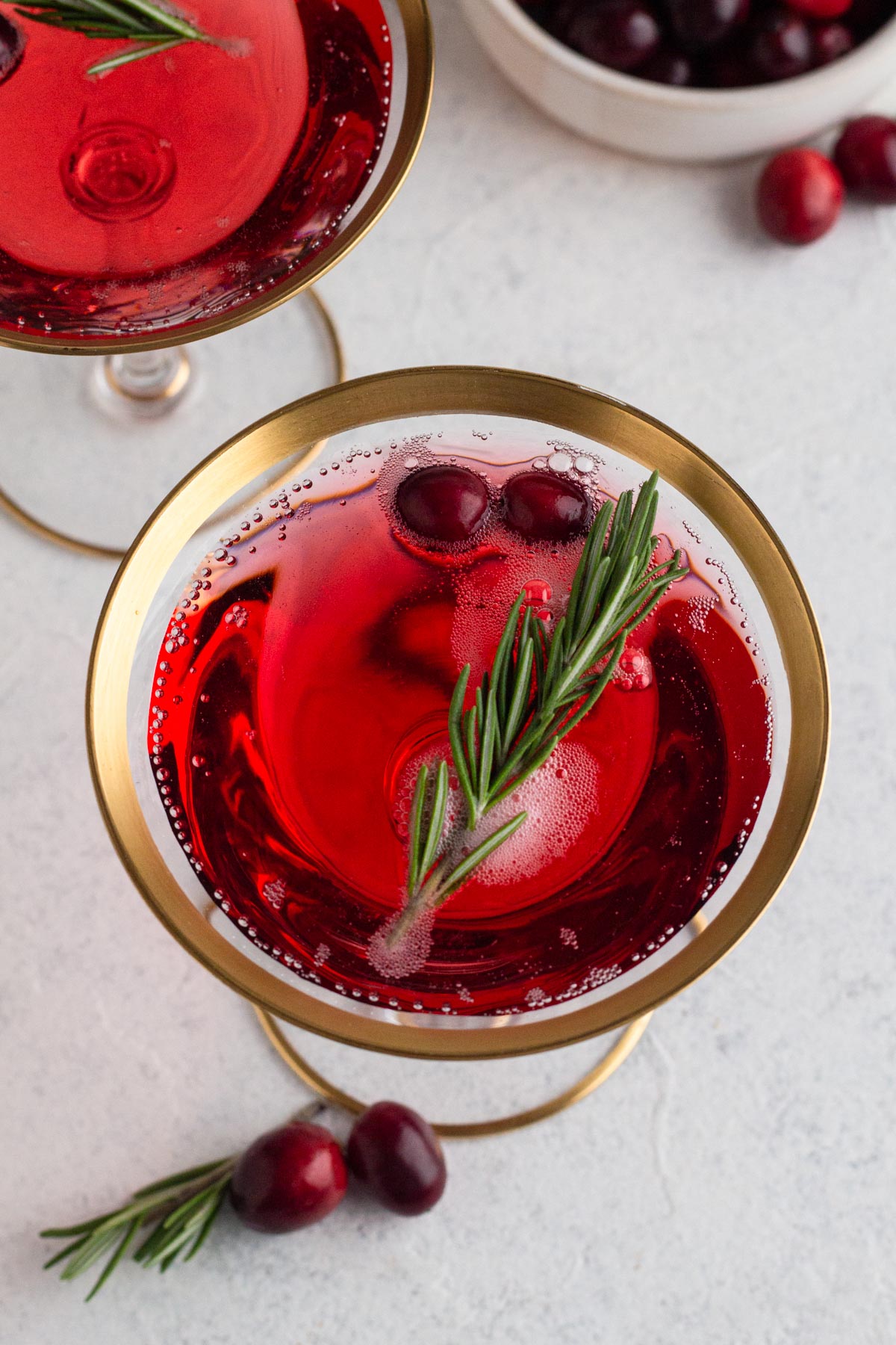 Champagne cocktail in a gold-rimmed coupe glass with fresh rosemary and cranberry garnish.