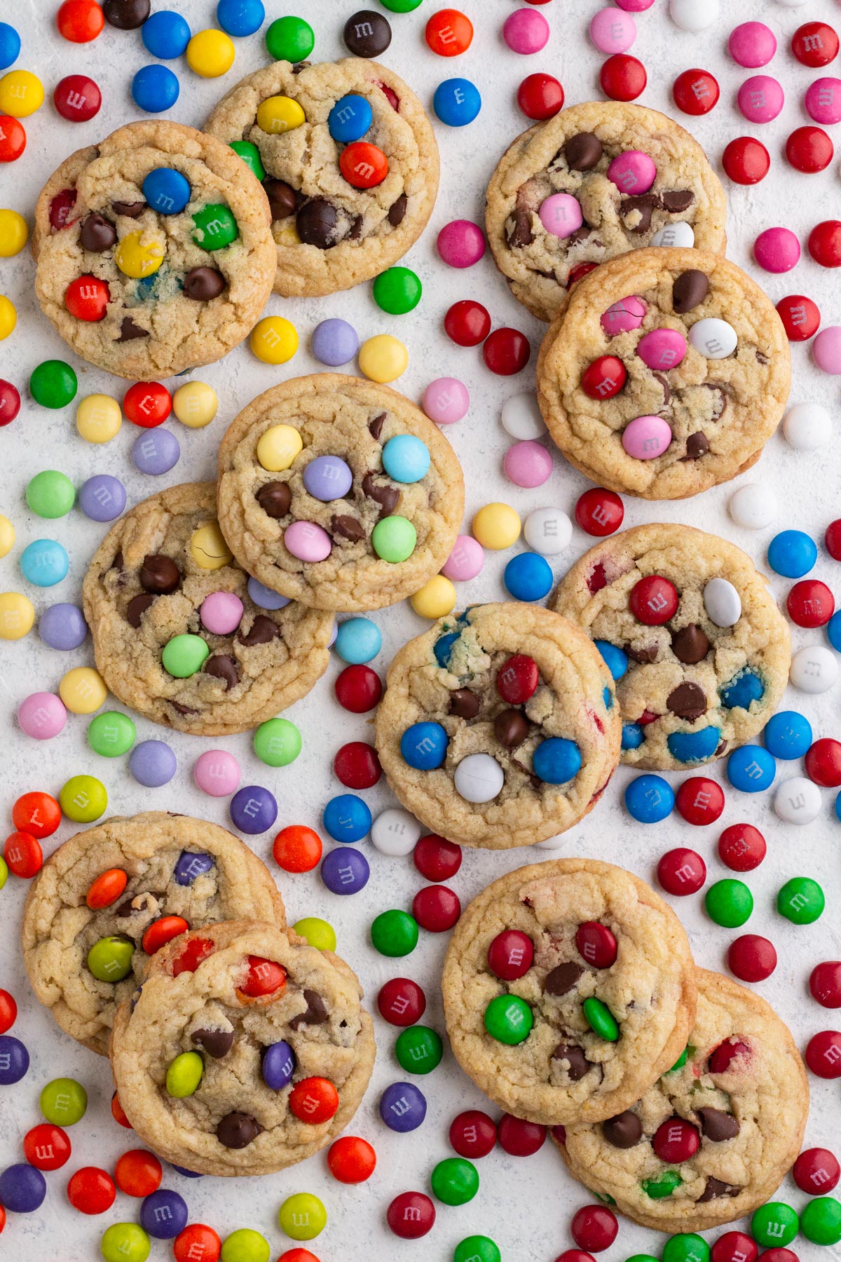Cookies with M&Ms and chocolate chips scattered on a white surface surrounded by assorted holiday M&Ms.