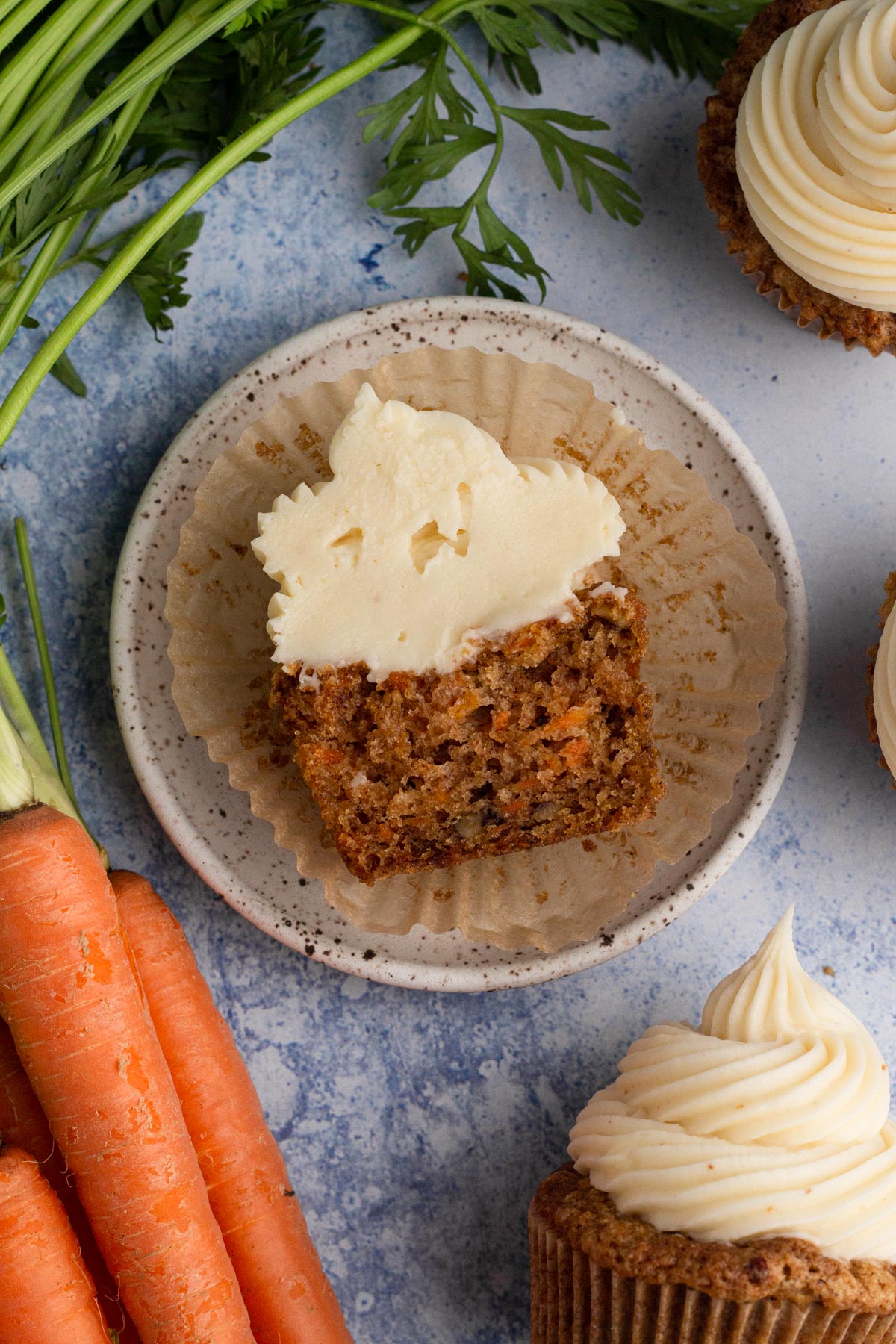 A frosted carrot cake cupcake sliced in half on a plate surrounded by carrots and more cupcakes.