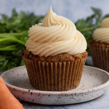 Close up view of a carrot cake cupcake topped with a swirl of cream cheese frosting on a speckled plate surrounded by carrots.