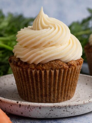 Close up view of a carrot cake cupcake topped with a swirl of cream cheese frosting on a speckled plate surrounded by carrots.