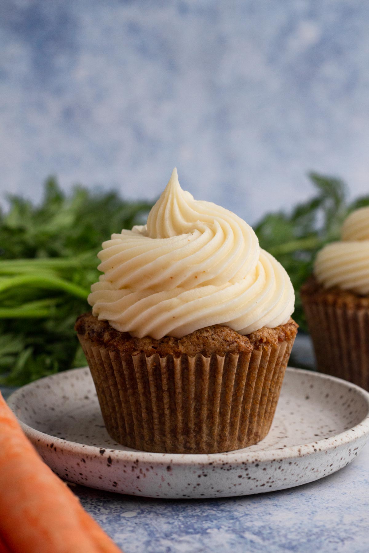 A carrot cake cupcake topped with a swirl of cream cheese frosting on a speckled plate surrounded by carrots.