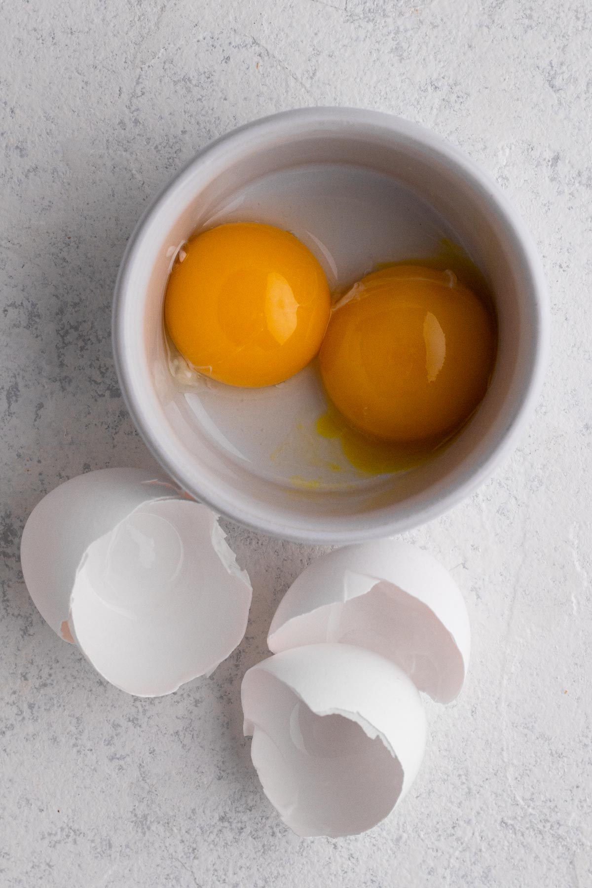 Bowls of egg yolks with cracked egg shells on a white surface.