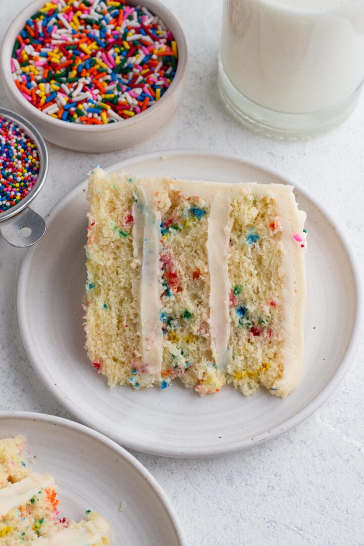 Slice of cake on a white plate surrounded by bowls of sprinkles and a bottle of milk.