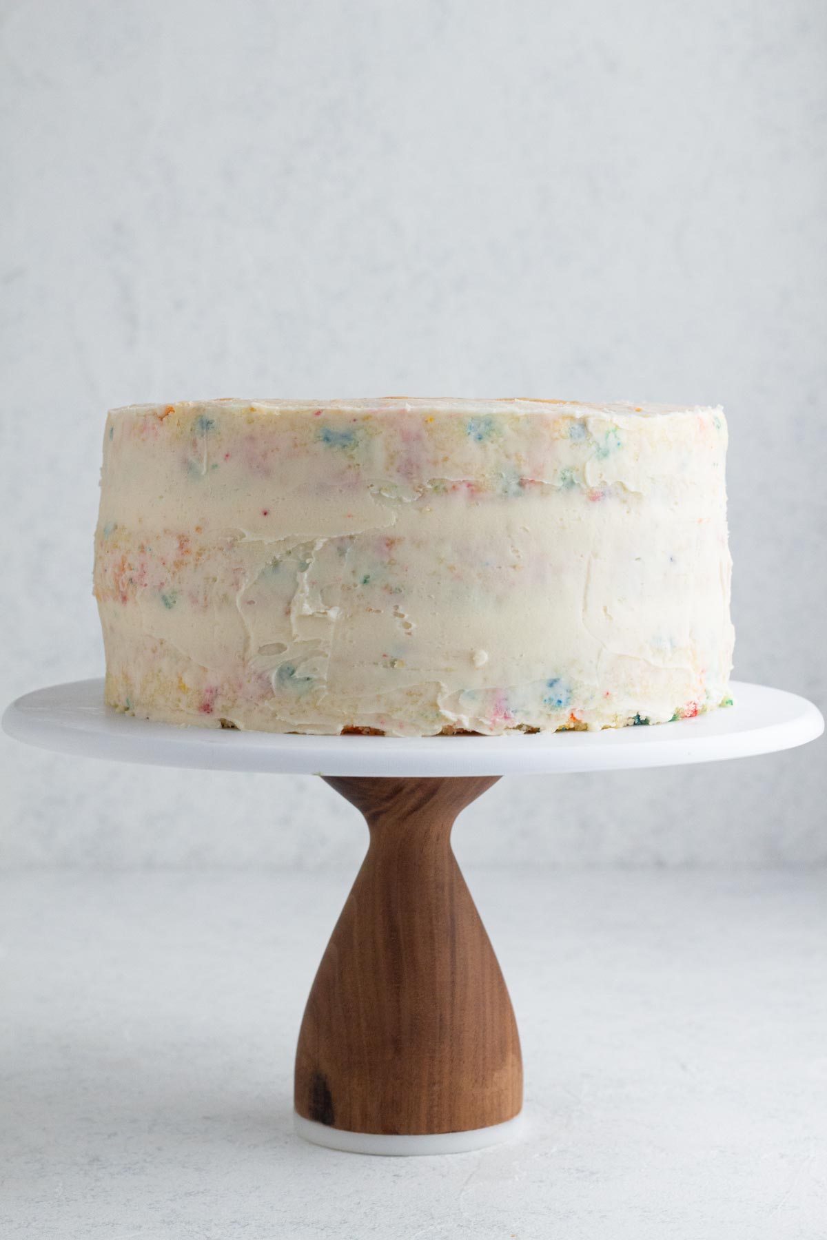 Crumb coat of frosting on funfetti layer cake on a wooden cake stand.