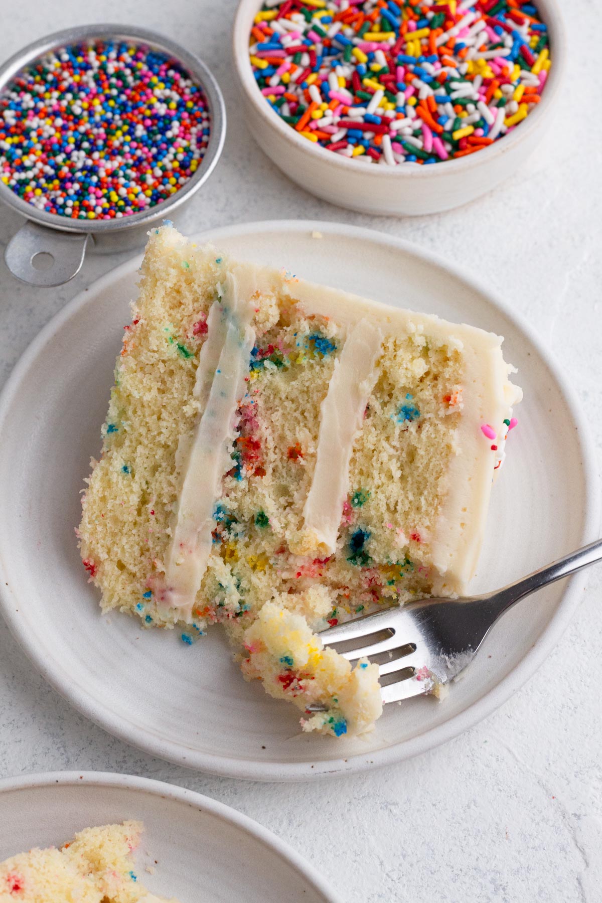Slice of cake with a forkful of cake on a white plate surrounded by bowls of sprinkles.