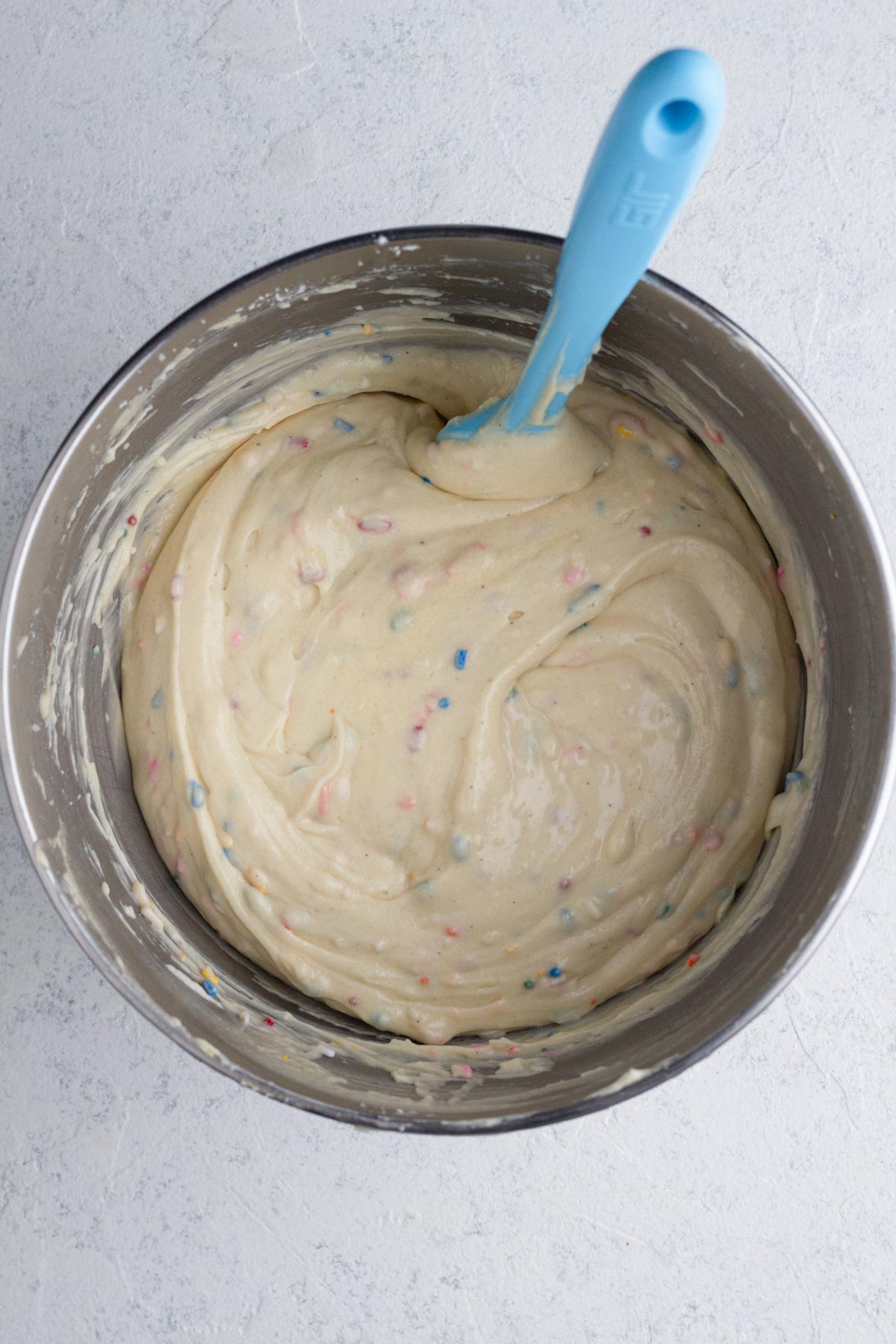 Cake batter with rainbow sprinkles in a metal bowl with a blue spatula.