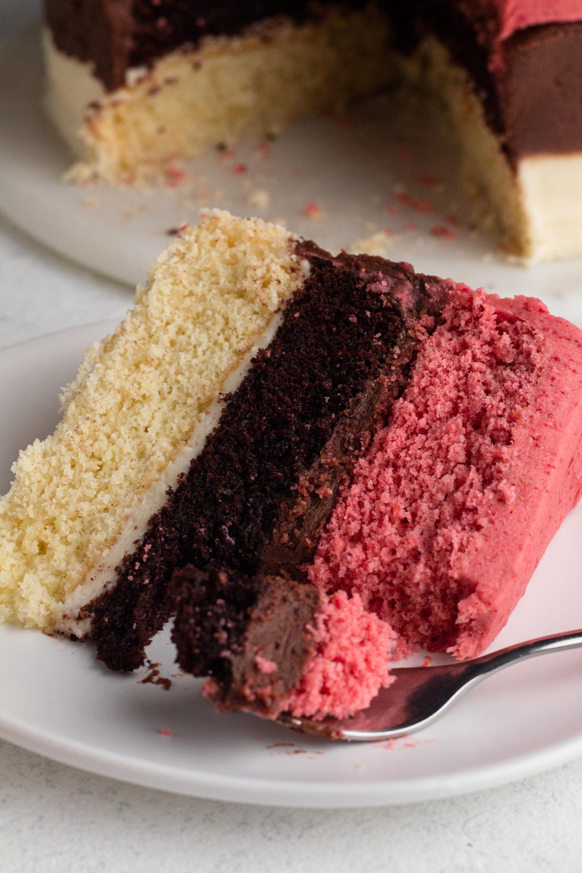 Close up view of a slice of neapolitan cake with a forkful of cake on a white cake plate.