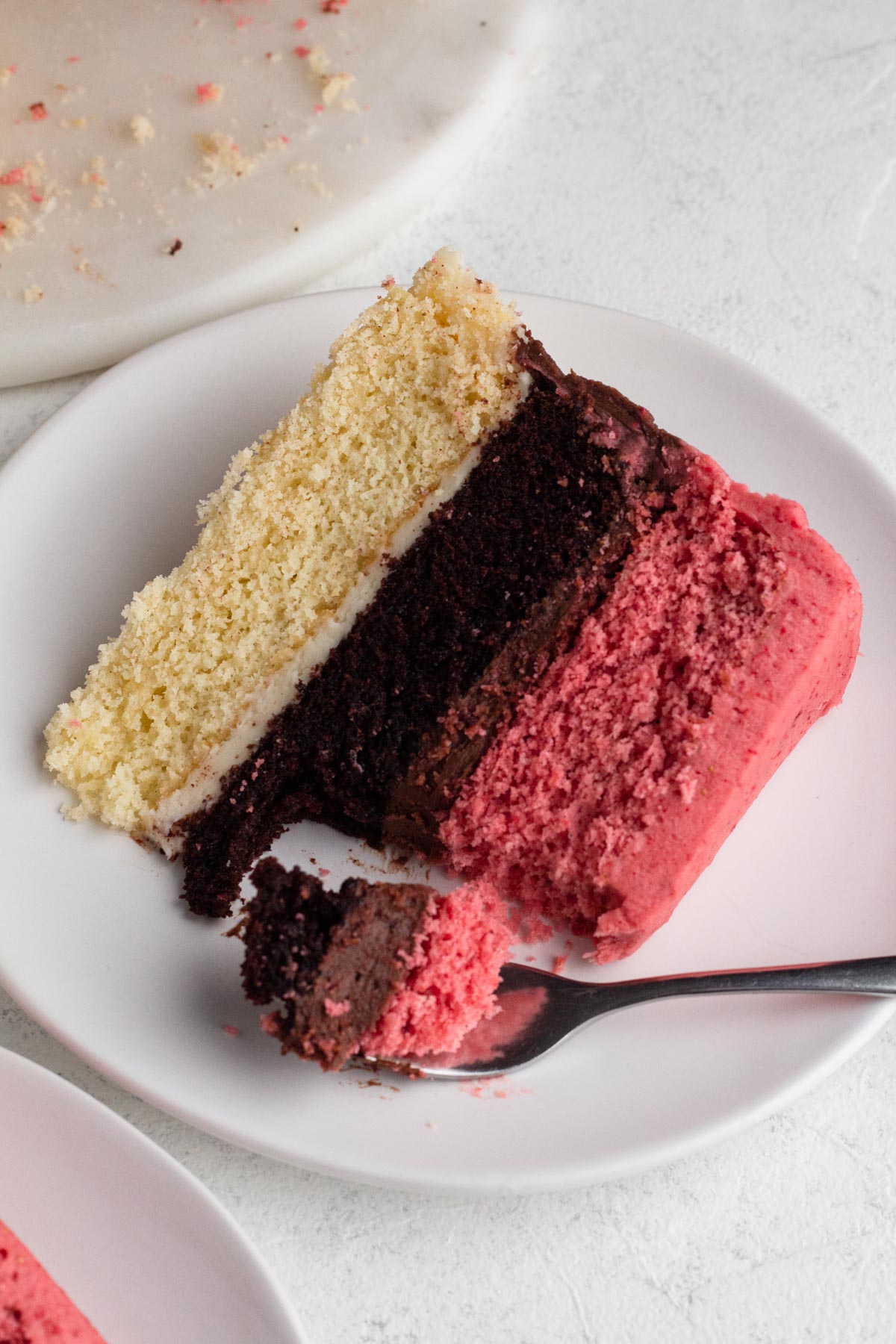 A slice of neapolitan cake with a forkful of cake on a white cake plate.