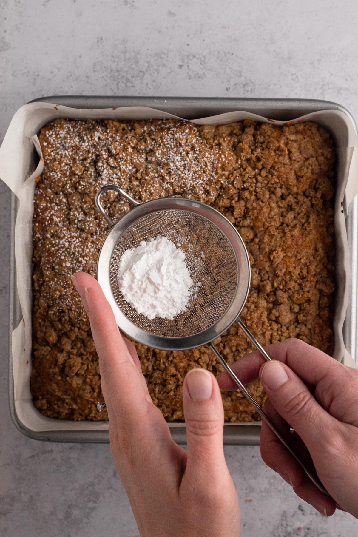 Hands using a small strainer to dust powdered sugar over a coffee cake in a square baking pan.