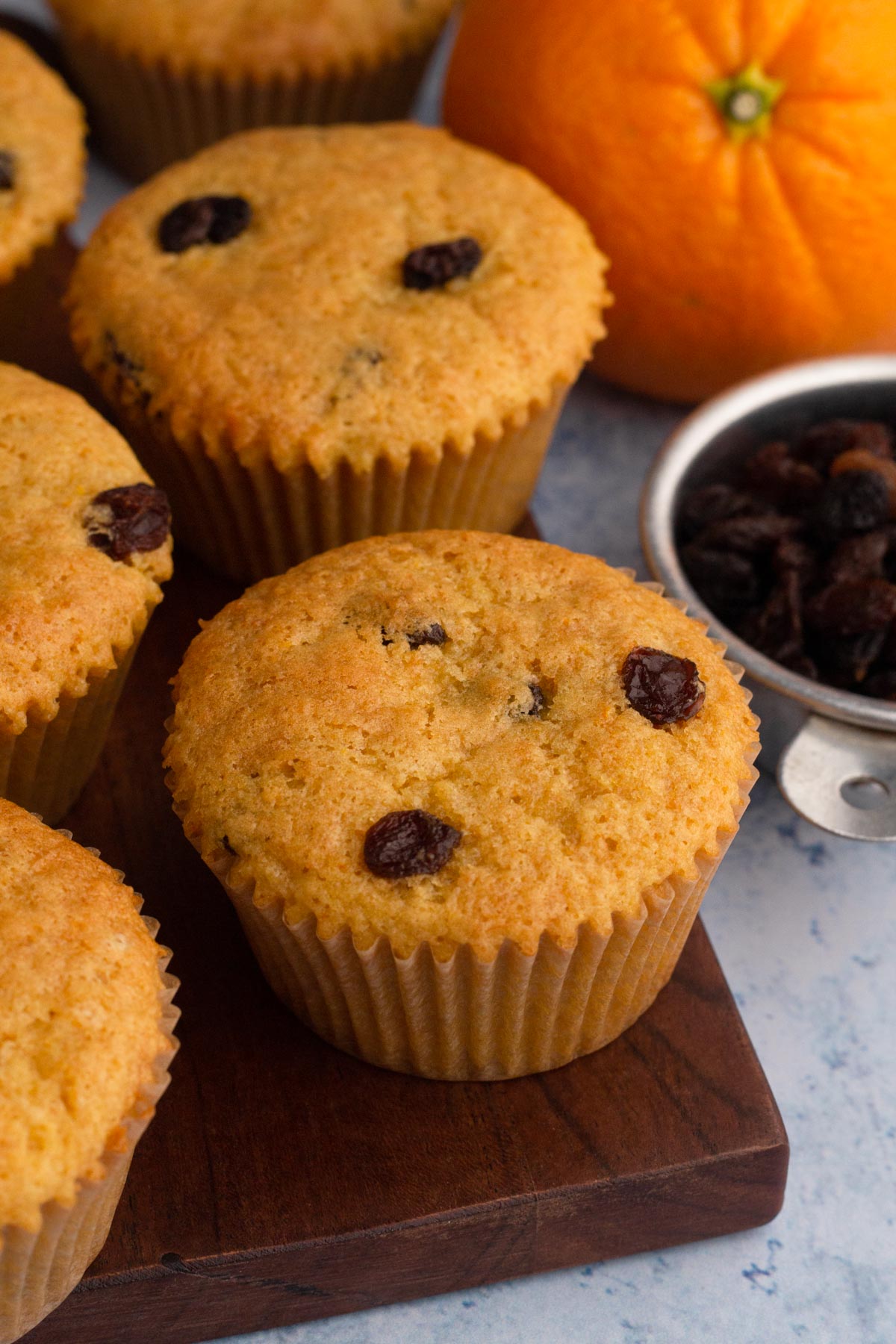 Muffins on a wooden cutting board with a whole orange and a bowl of raisins.