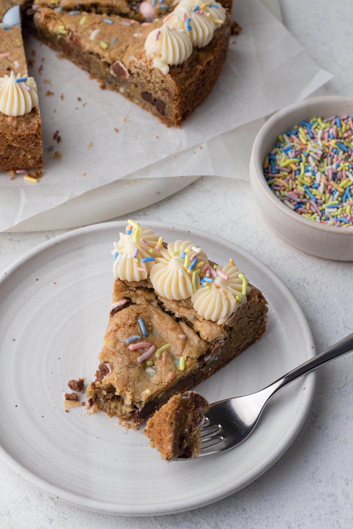 A slice of cookie cake decorated with vanilla frosting and pastel sprinkles on a white plate.