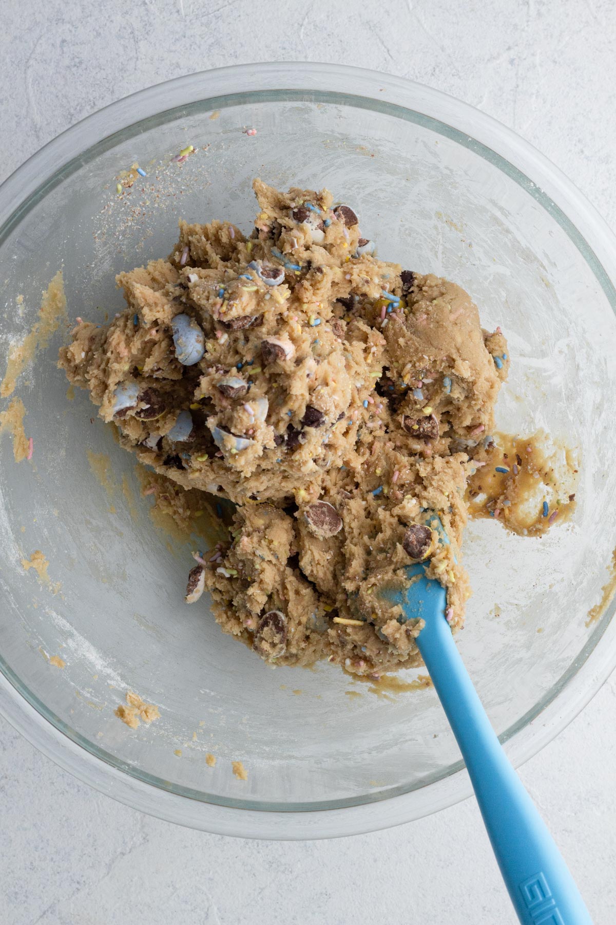 Cookie dough with candy mix-ins and a blue spatula in a glass mixing bowl.