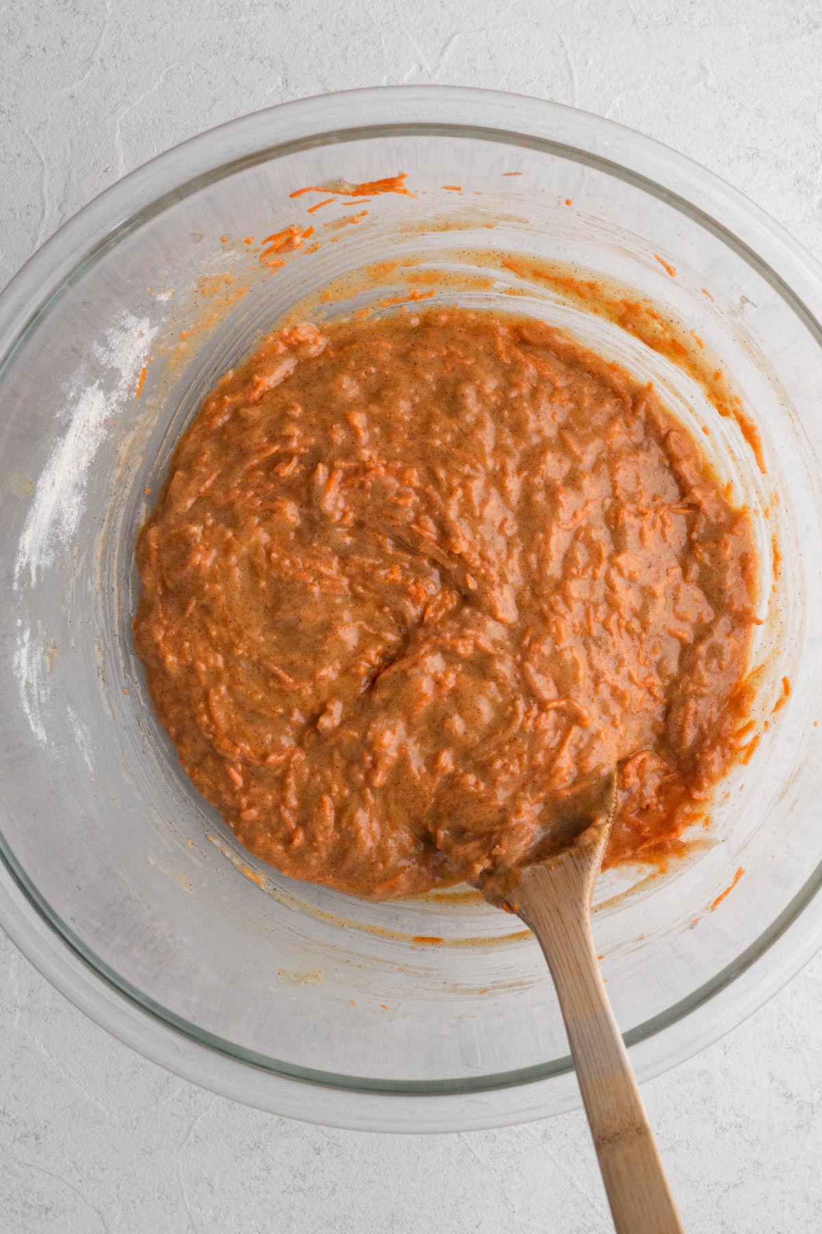 Cake batter with shredded carrots in a glass mixing bowl with a wooden spoon.