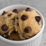 Close up view of scoops of chocolate chip cookie dough in a white ramekin on a gray surface.
