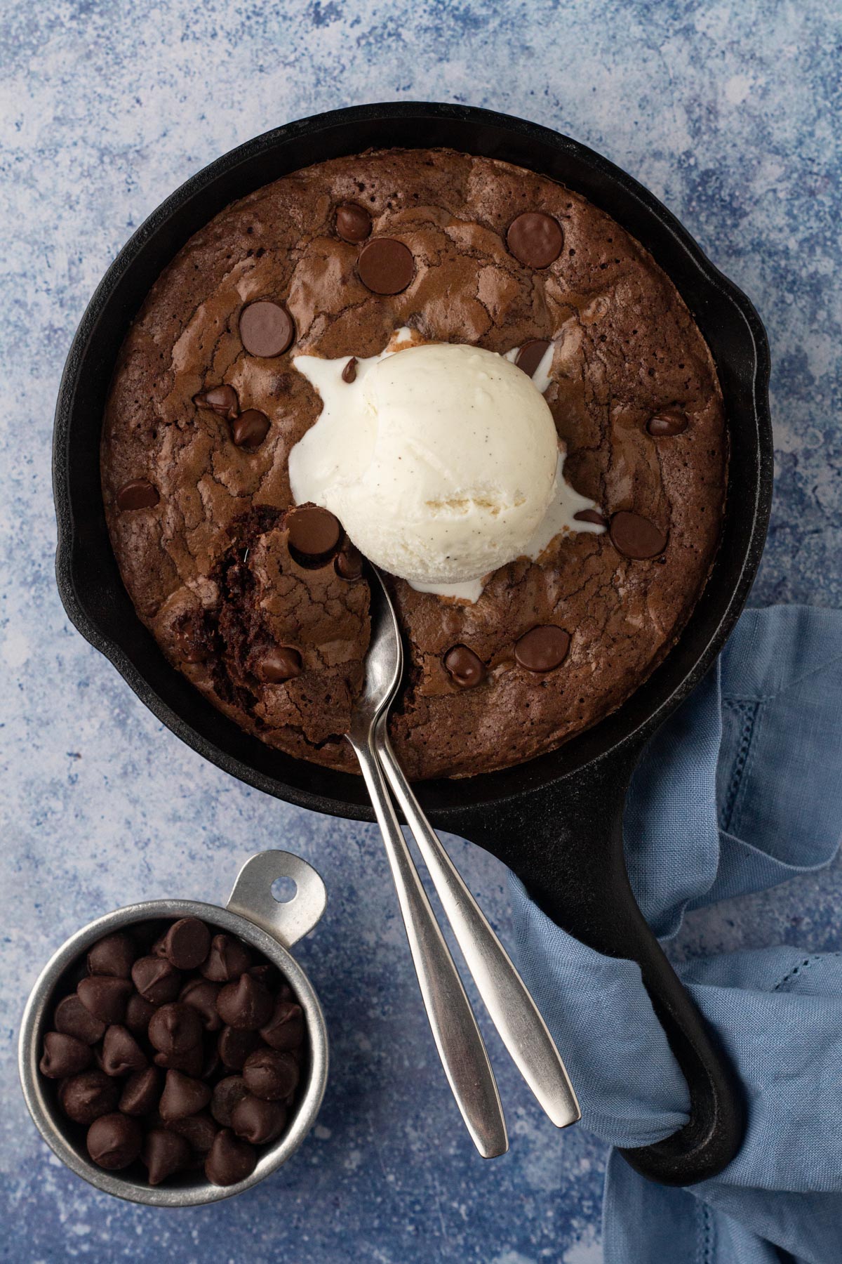 Mini skillet brownie with a scoop of vanilla ice cream and two spoons on a blue surface.