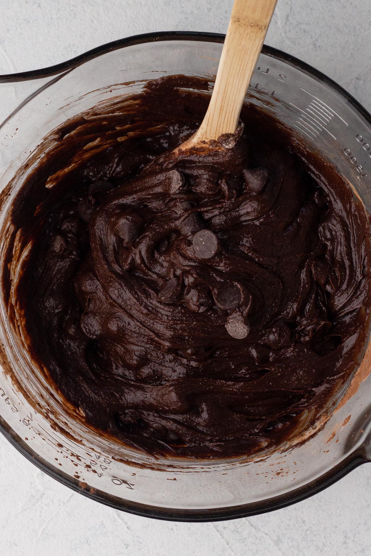 Brownie batter with chocolate chips in a glass batter bowl with a wooden spoon.