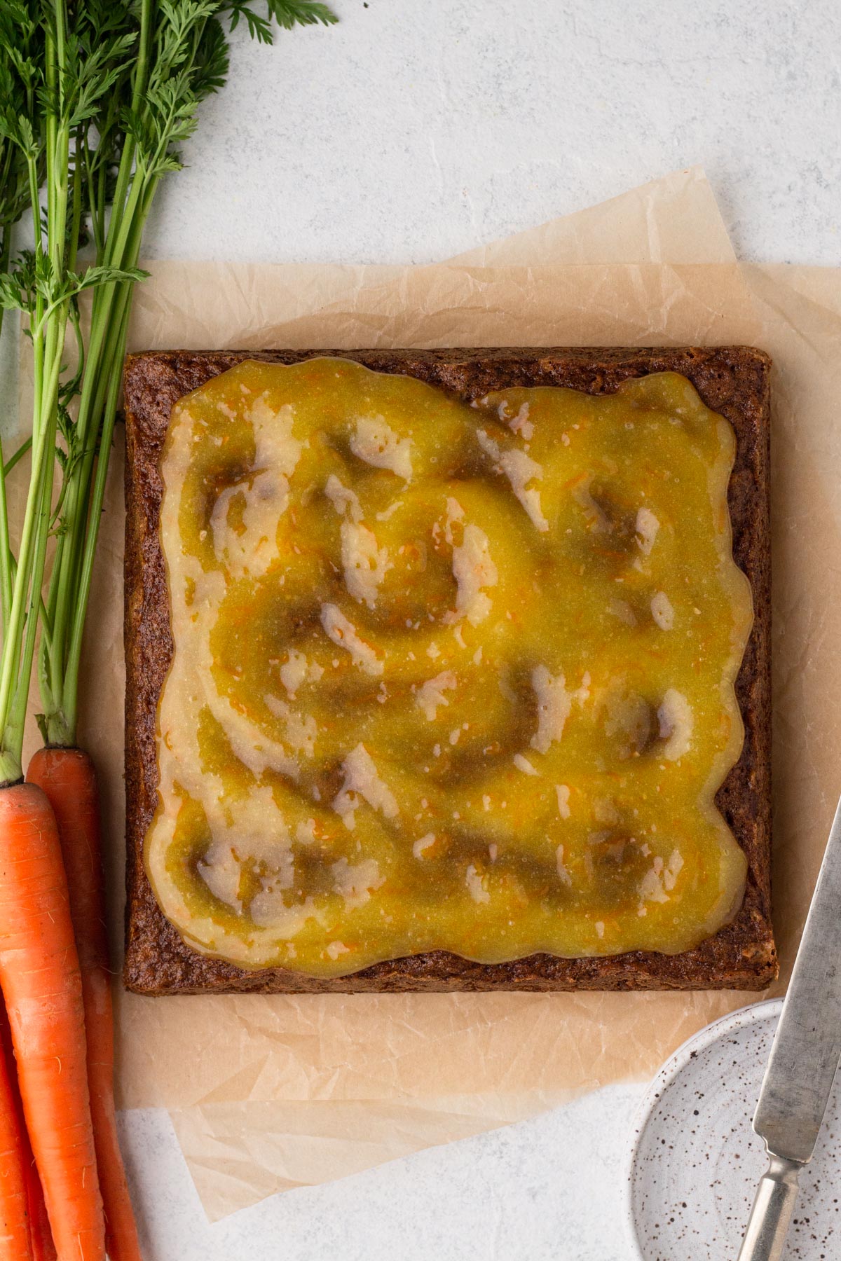 Square carrot cake with orange glaze on parchment paper with fresh carrots.