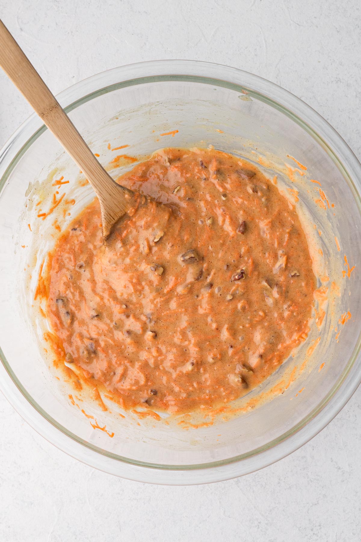 Cake batter with carrots and pecans in a glass mixing bowl with a wooden spoon.
