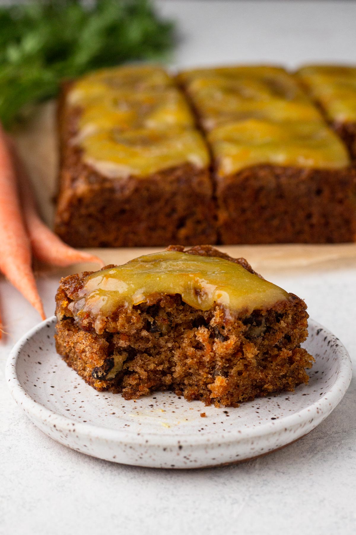 A square slice of carrot cake topped with orange glaze on a white plate with the other cake slices and fresh carrots in the background.