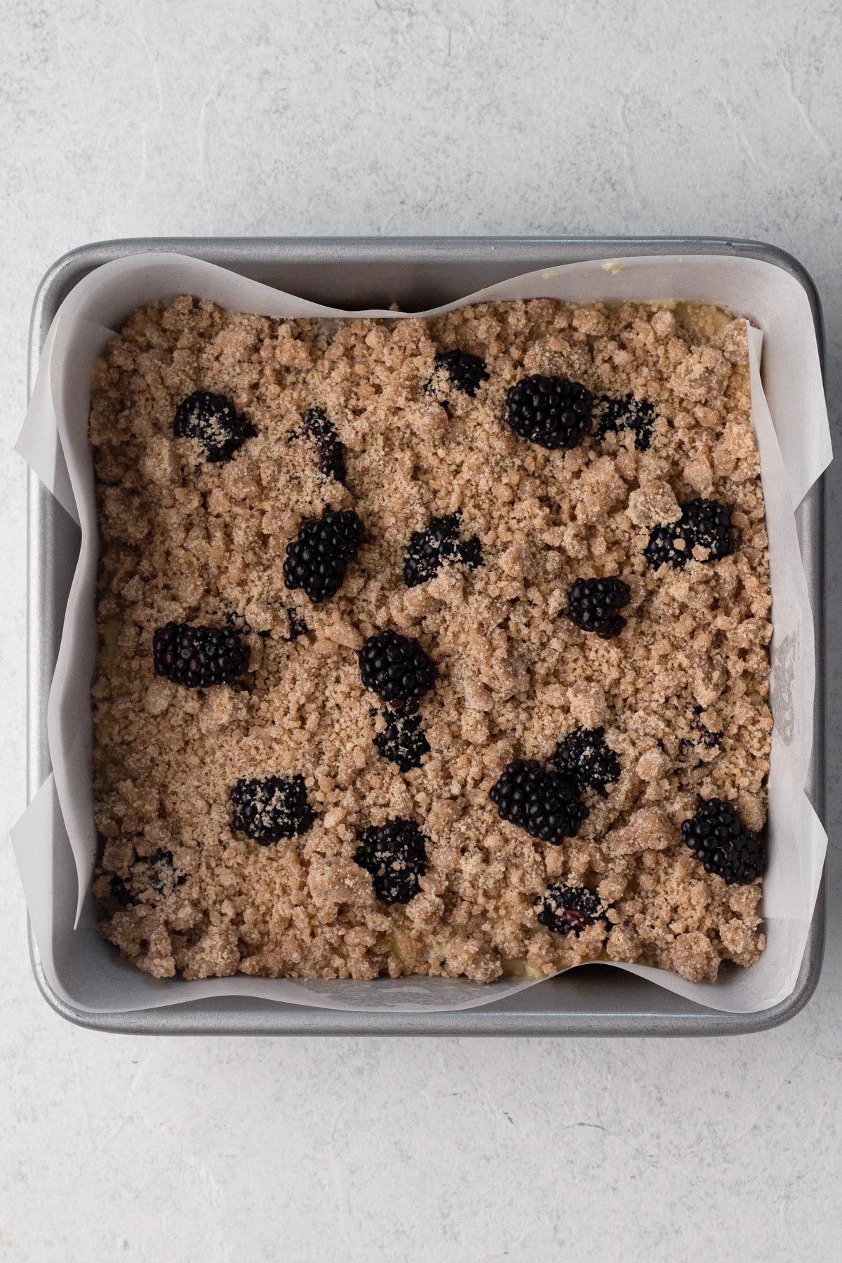 Cake batter topped with crumb topping and blackberries in a square metal baking pan.