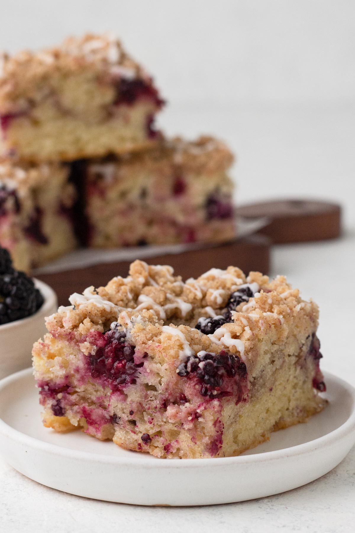 A square slice of coffee cake with blackberries and a drizzle of glaze on a white plate.