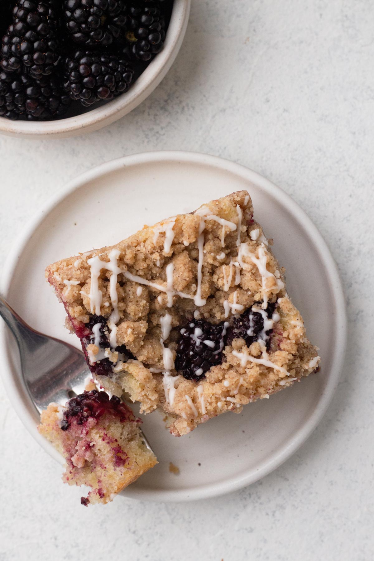 Overhead view of a square slice of coffee cake with blackberries and a drizzle of glaze on a white plate.