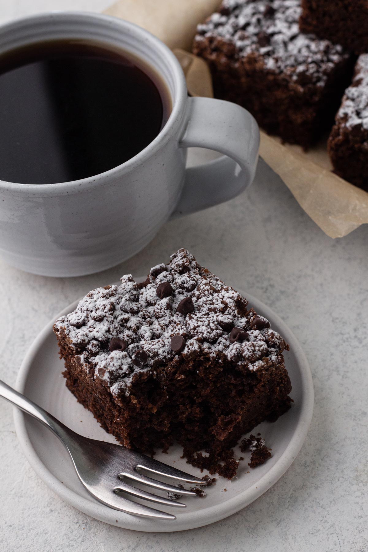 Angled view of a bitten slice of chocolate crumb cake on a white plate with a fork next to a mug of coffee.