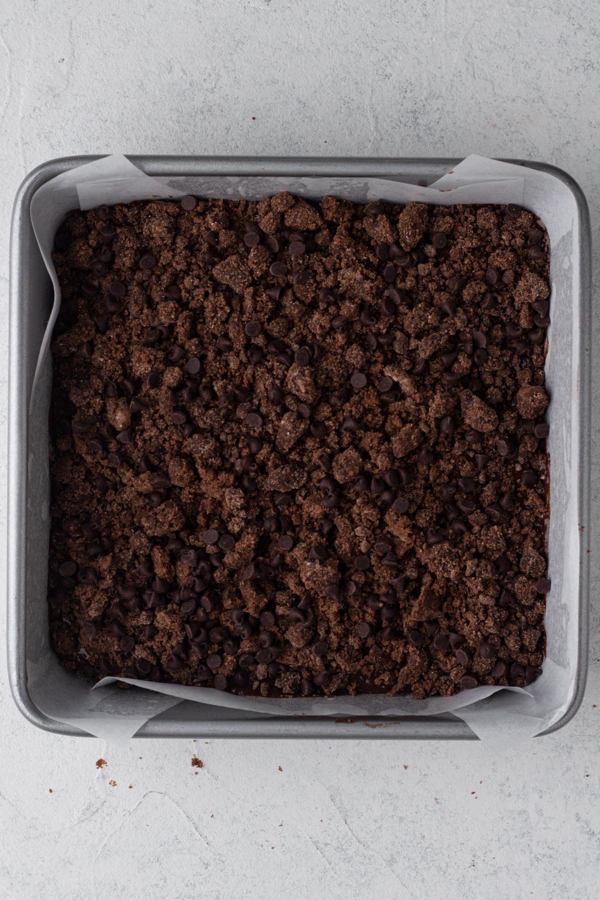 Chocolate cake batter layered with chocolate crumbs in a square metal baking pan lined with parchment paper.