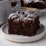 A slice of chocolate crumb cake on a white plate next to a mug of coffee.