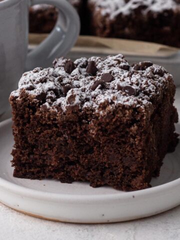 A slice of chocolate crumb cake on a white plate next to a mug of coffee.