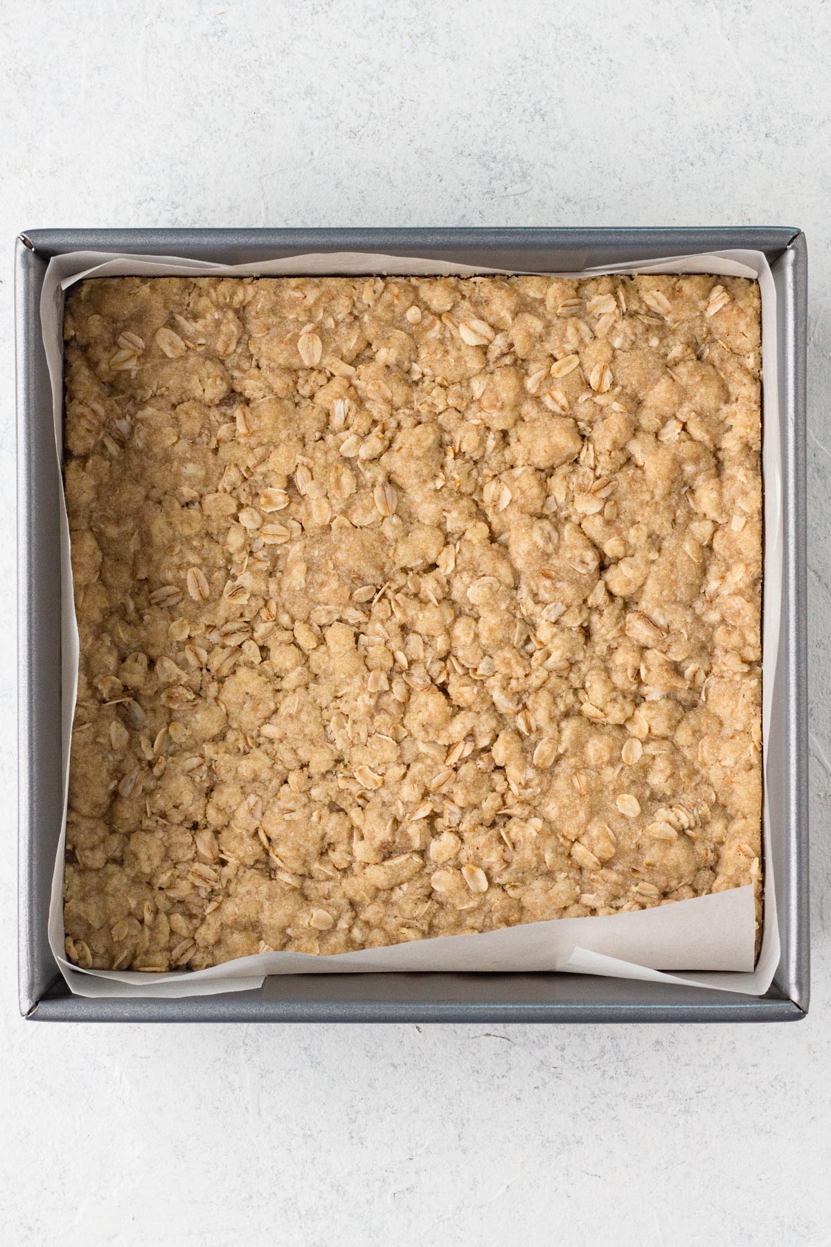 Parbaked crumble crust in a square metal baking pan lined with parchment paper.