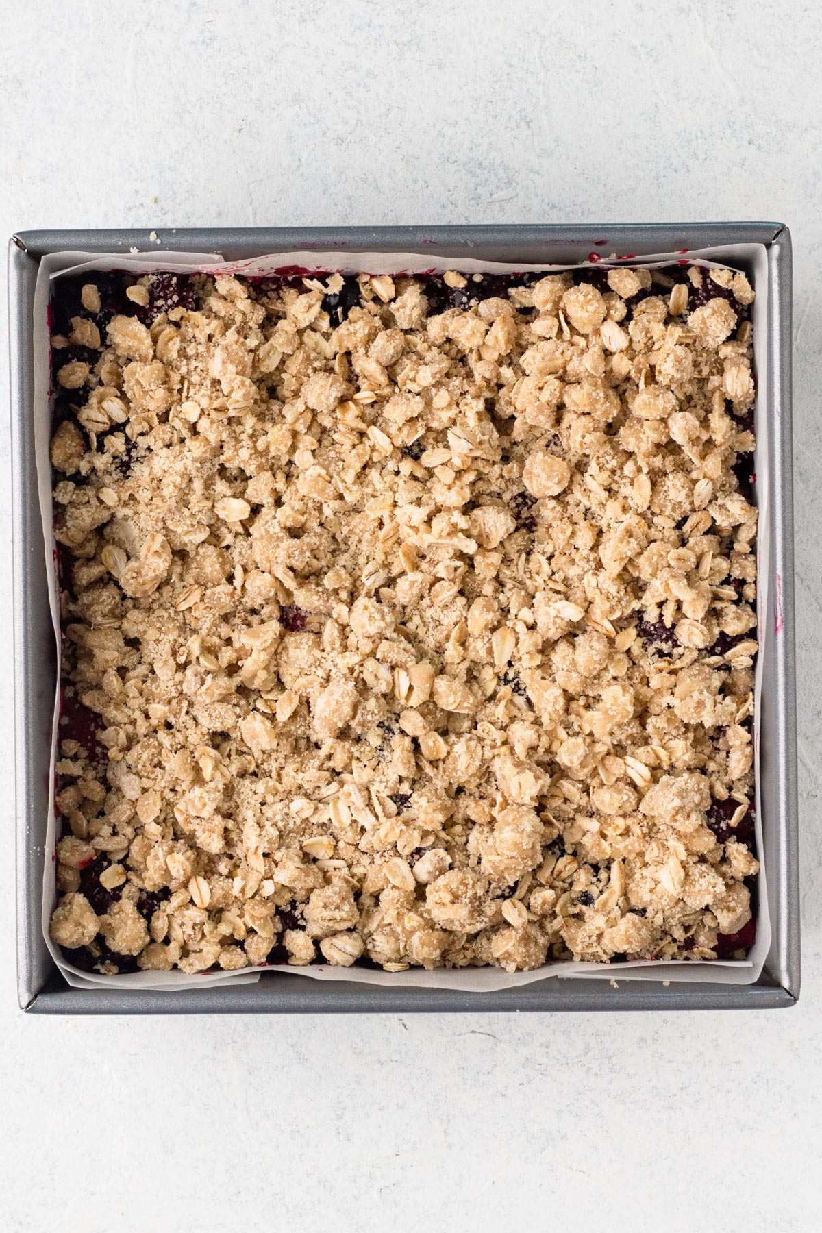 Parbaked crust topped with fruit filling and crumble topping in a square metal baking pan lined with parchment paper.