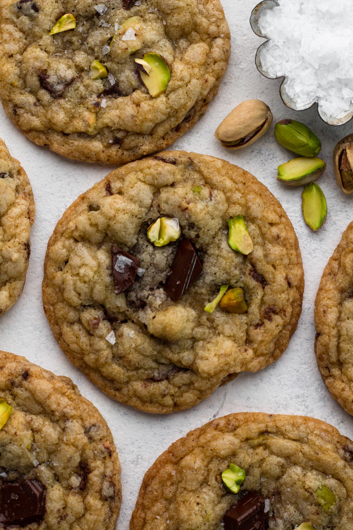 Cookies with bits of chocolate and chopped pistachios on a white surface with a dish of flaky sea salt.