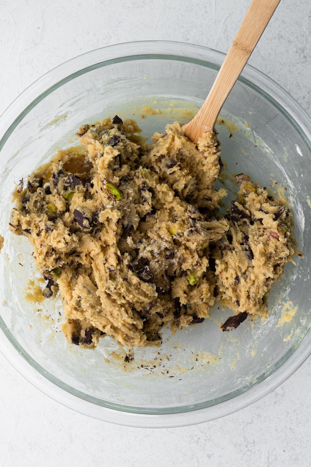 Cookie dough with bits of chocolate and pistachios in a glass mixing bowl with a wooden spoon.