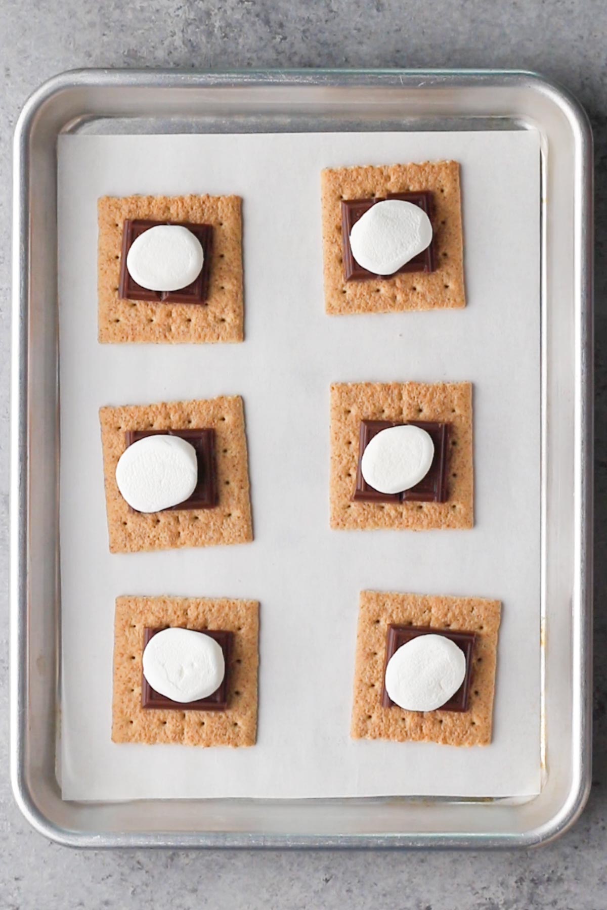 Graham crackers topped with squares of chocolate and marshmallows on a baking sheet.