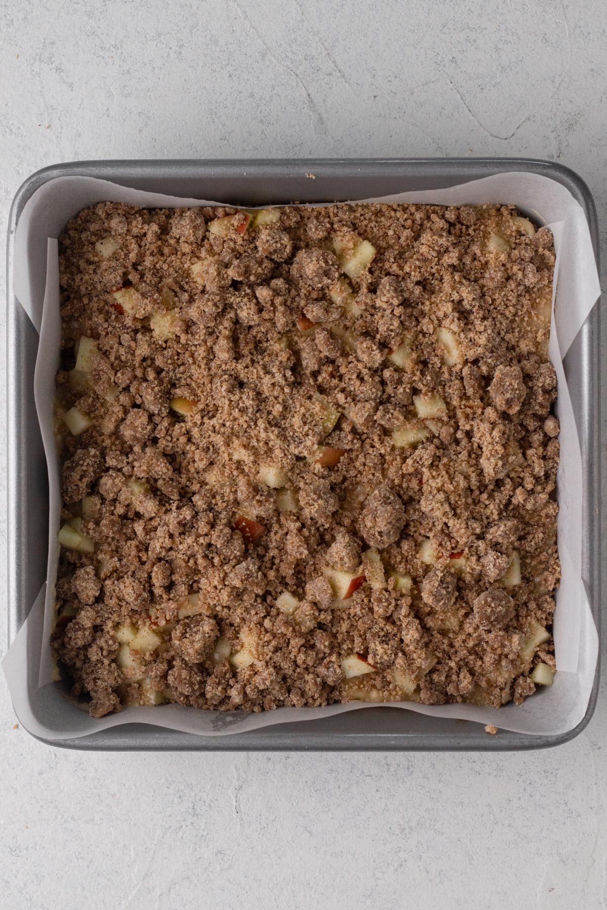Cake batter layered with chopped apples and crumb topping in a square baking pan.