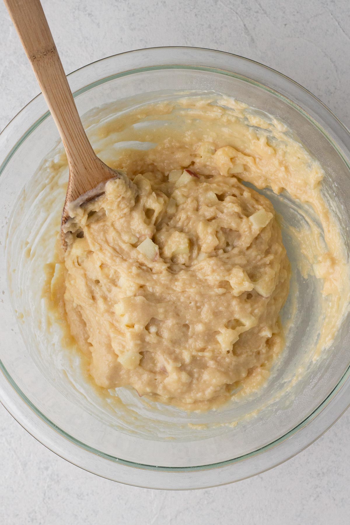 Cake batter with apples in a glass mixing bowl with a wooden spoon.