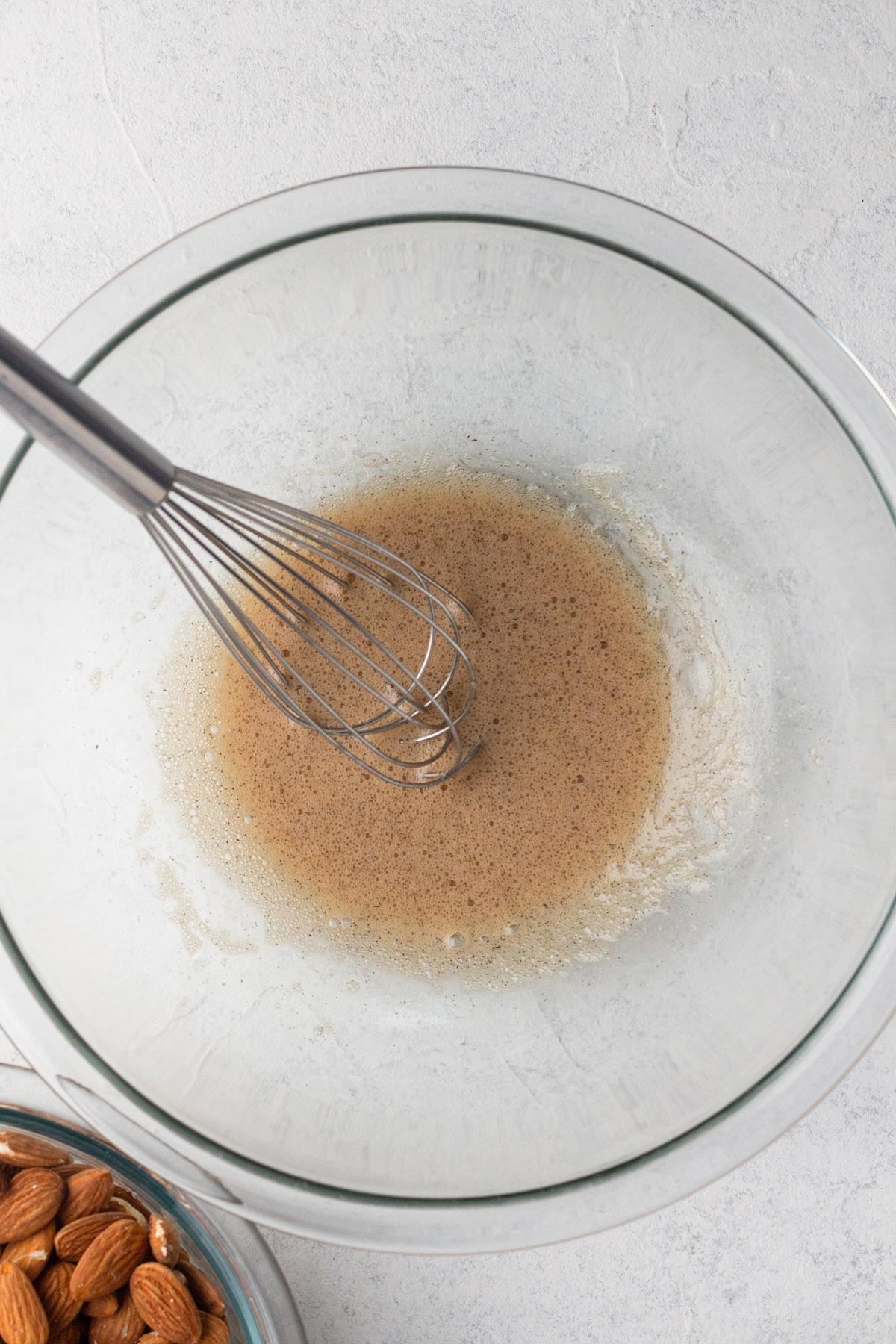 Frothy egg white with vanilla bean paste in a glass mixing bowl with a whisk.