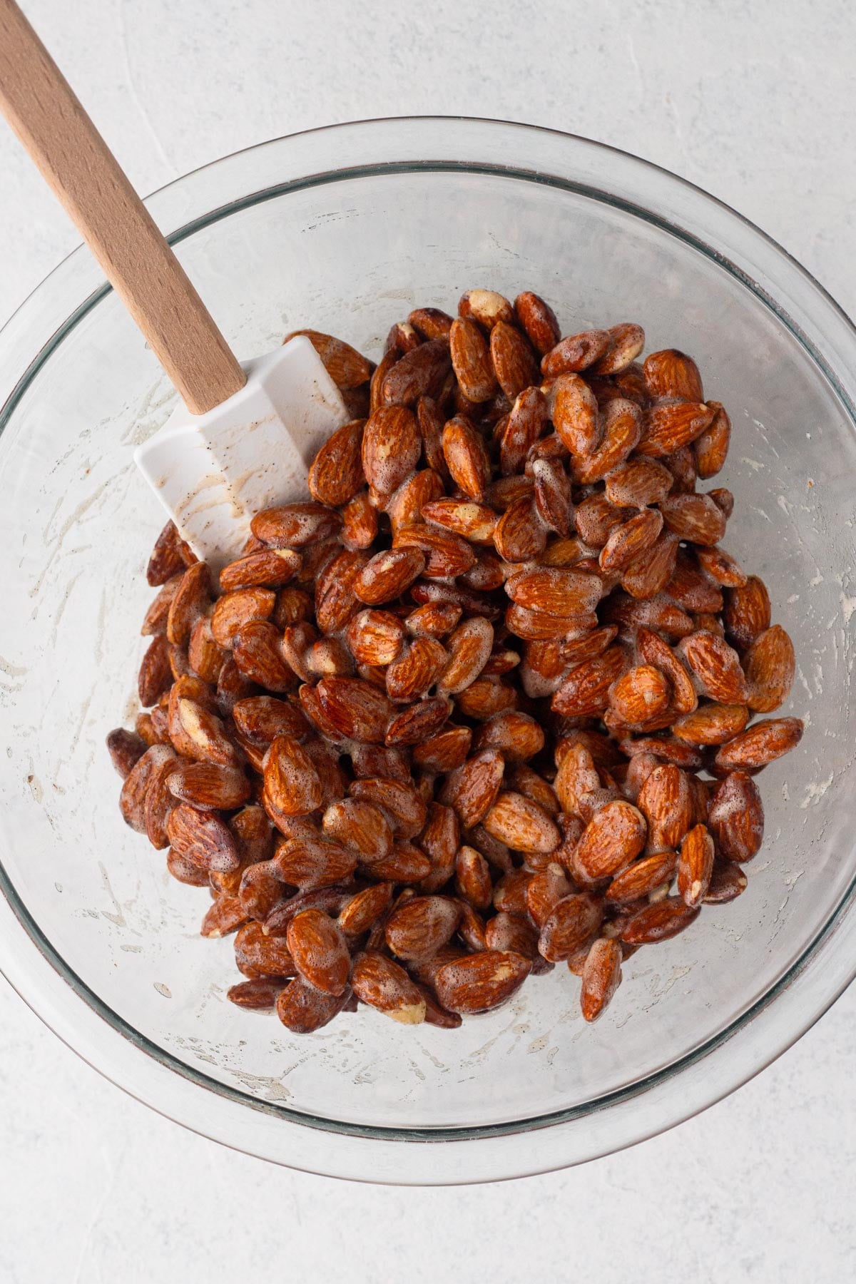 Raw almonds coated with egg white in a glass mixing bowl with a spatula.