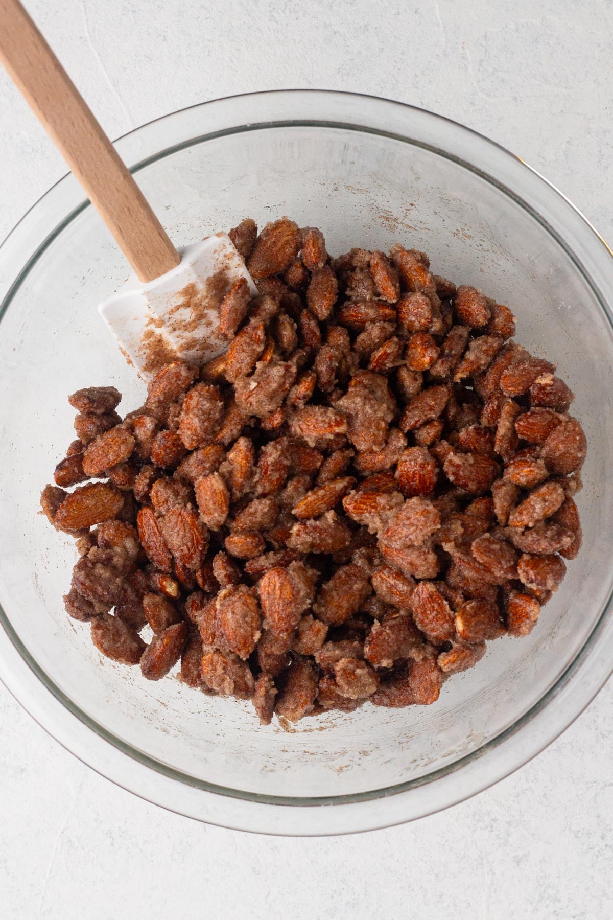 Raw almonds coated with egg white and cinnamon-sugar in a glass mixing bowl with a spatula.